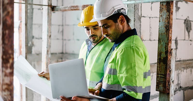 Two-construction-workers-at-building-site-looking-at-blue-print-and-laptop